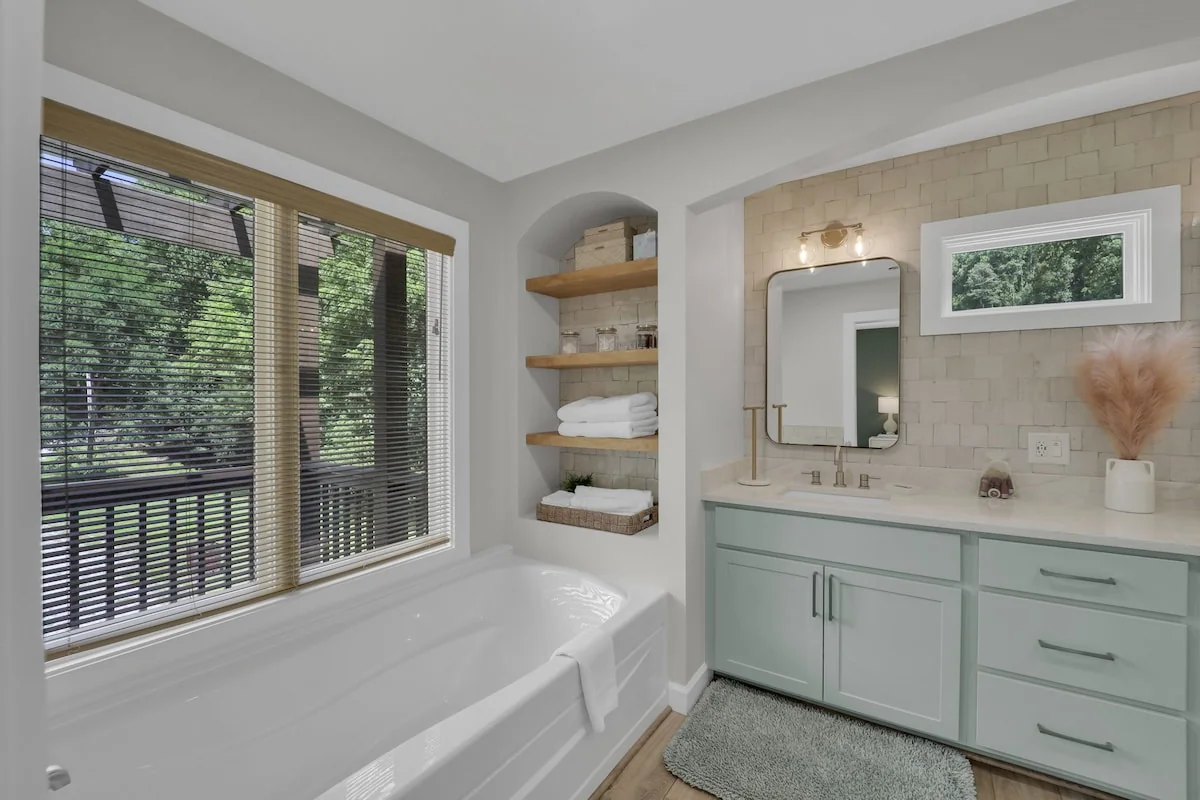Soaking tub under a wooded-view window, pale green vanity, mirror, and towel shelves.